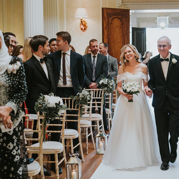 Father of the bride in black tie walks the bride in off the shoulder wedding dress down the aisle 