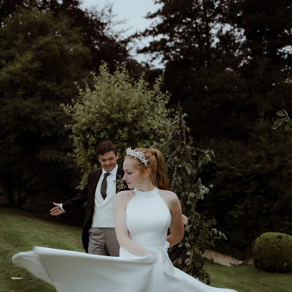 Bride spins around in a halter neck pleated skirt wedding dress and pearl crown with groom watching in the background 