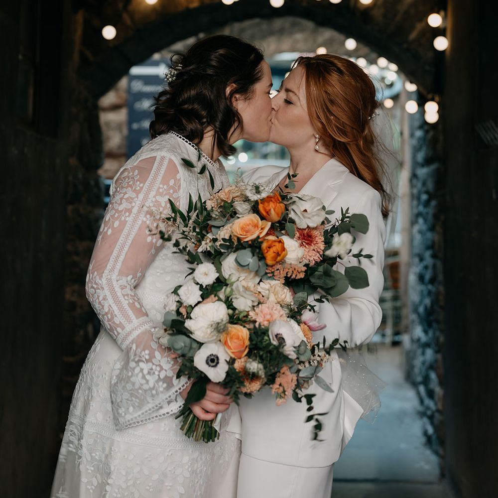 Brides kiss on their wedding day as they hold white and orange floral bouquets 