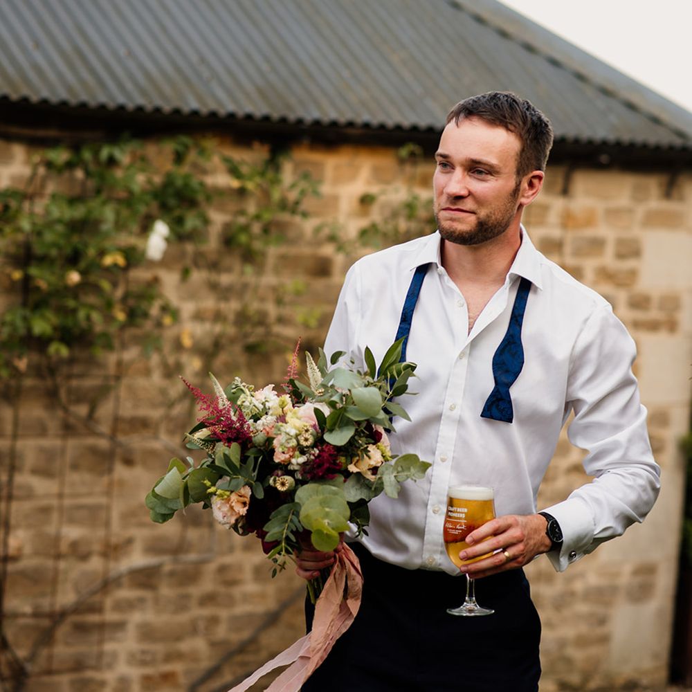 Groom in white shirt and undone blue bow tie holding glass of beer and the red and white bridal bouquet