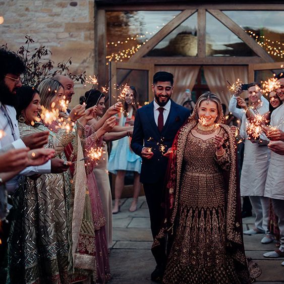 Bride wearing brightly coloured saree finished with gold embellishment walks through sparkler exit with her groom in blue three piece suit