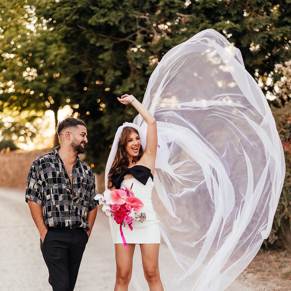 Bride wears mini dress with black bow and holds brightly coloured floral bouquet as she throws out her veil beside her 