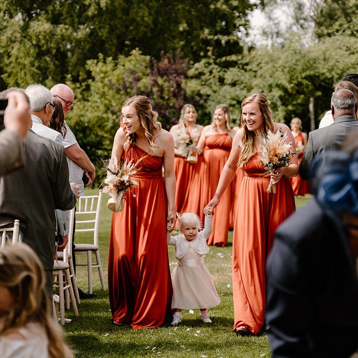 Bridesmaids in orange dresses walk down the aisle with a flower girl in a pale pink tulle dress