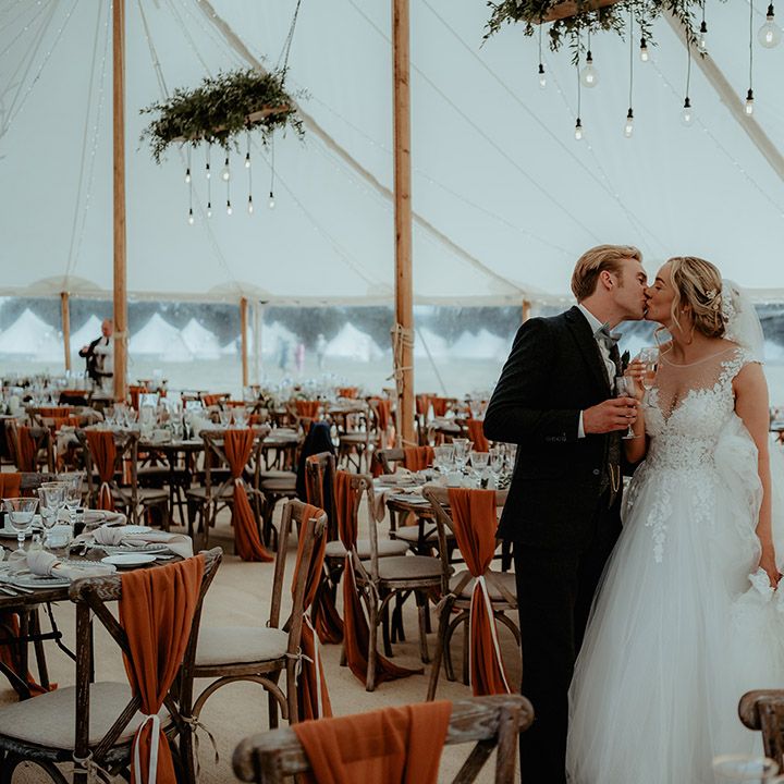 Bride and groom share a kiss in their marquee wedding venue with burnt orange decor, foliage and festoon lighting 