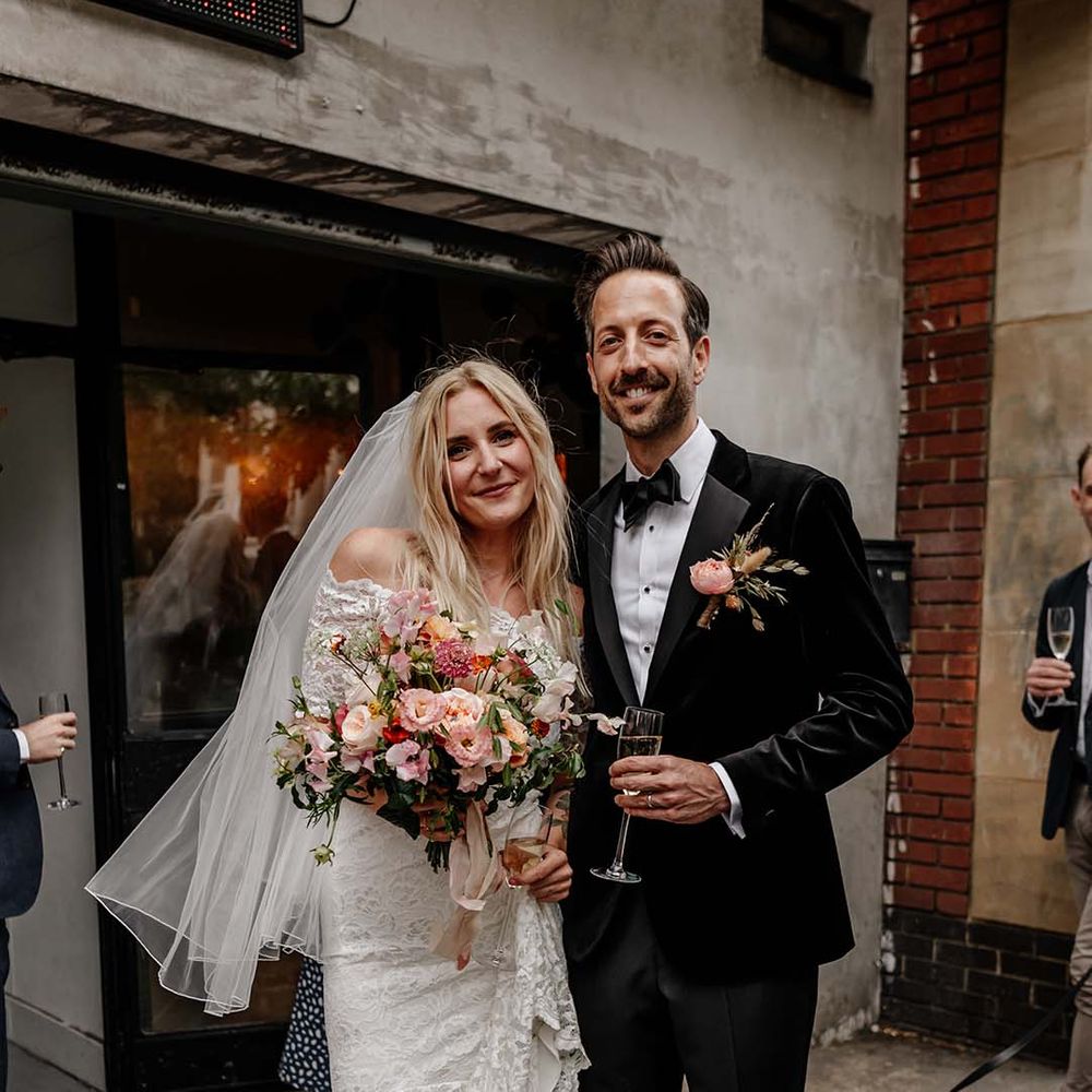 Bride in lace boho two piece, colourful mixed floral bouquet, gold strappy heels and short veil and groom in black tux with bow tie and boutonniere