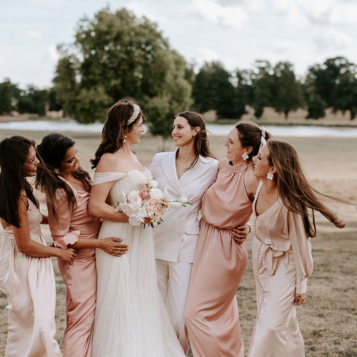 The bride in a white bridal suit smiling with the bride in an off the shoulder wedding dress with bridesmaids in pink and champagne coloured dresses 