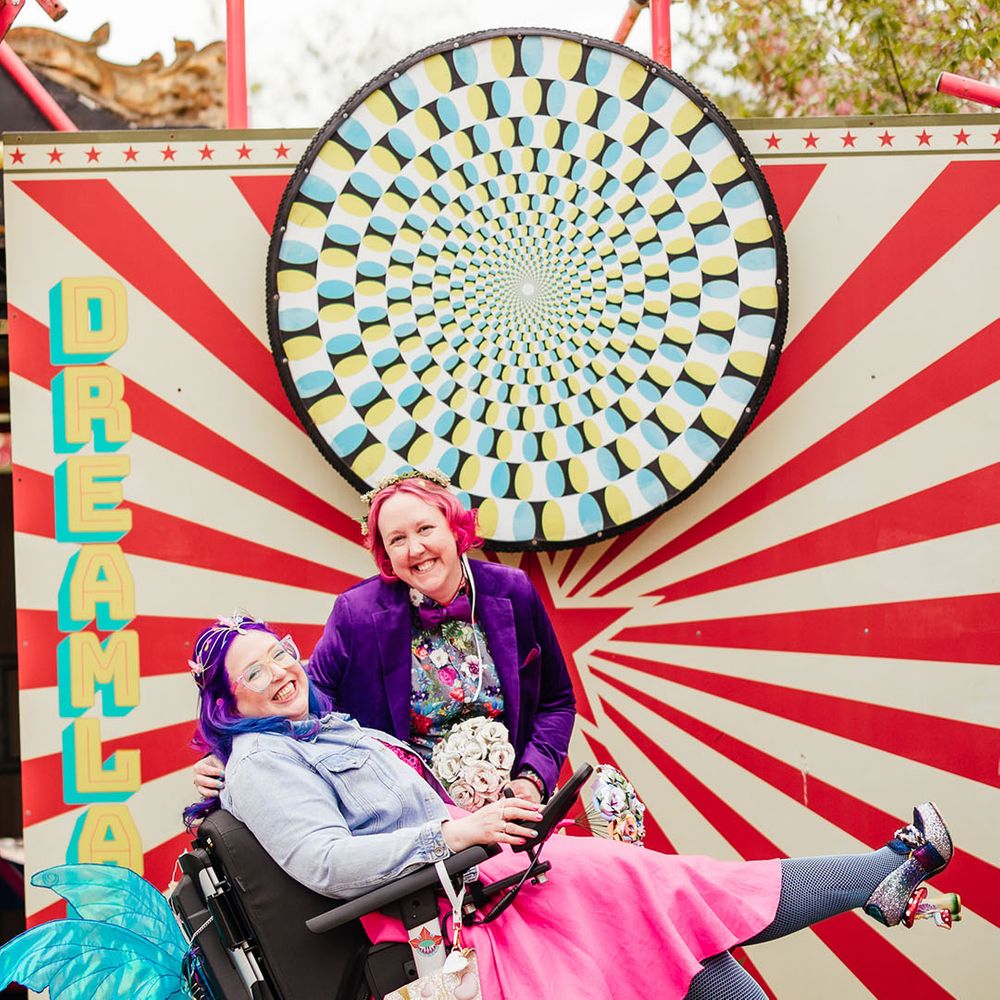 Bride in purple velvet blazer and black suit trousers with purple bowtie, pink patterned pocket square and colourful patterned shirt and bride in wheelchair wearing personalised denim jacket, pink wedding dress and blue tights posing in front of kaleidoscope sign at theme park wedding 