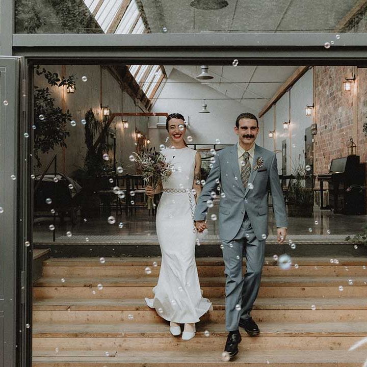 Lisa in pre loved fitted wedding dress walks alongside Gabriel in powder blue Gucci suit through bubbles 
