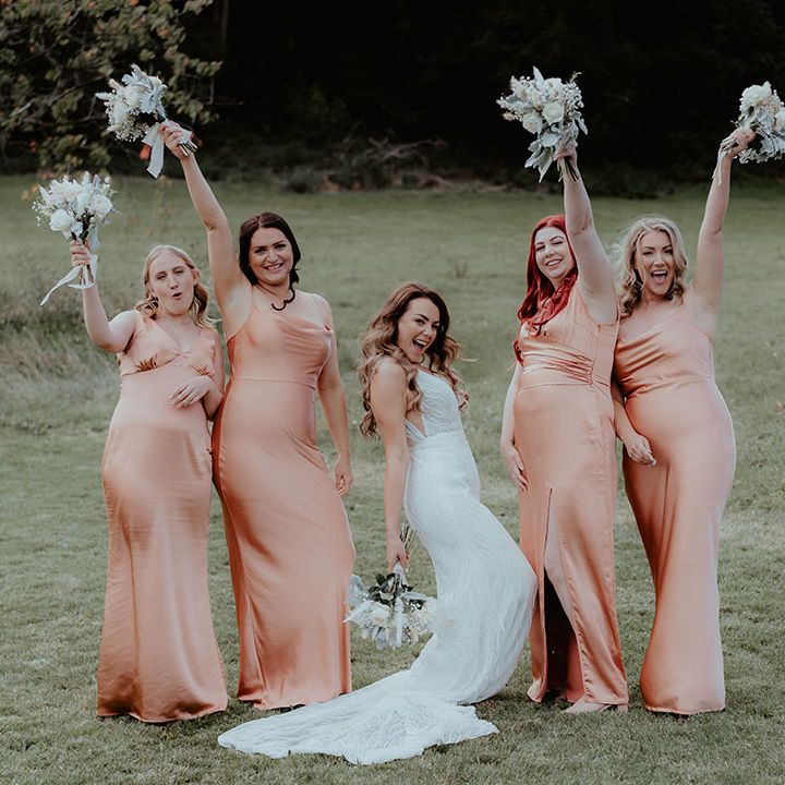 Bridesmaids in satin peach birdemaid dresses raise their white rose and gypsophila bouquets in the air with the bride 