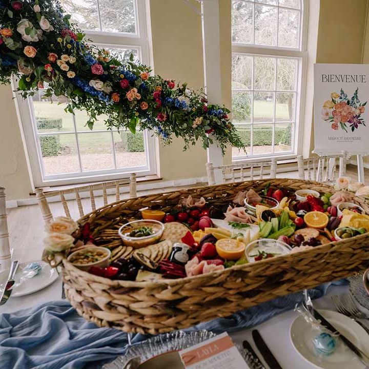 Large colourful wedding charcuterie board in wicker basket on classic wedding tablescape with white wedding tablecloth and cornflower blue wedding table runner