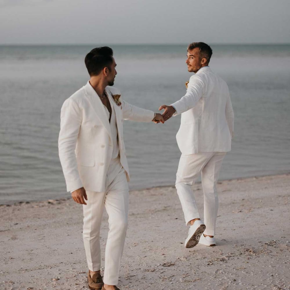 Grooms in complimenting double breasted white wedding suits holding hands doing couples photoshoot on the beach at Mexico destination wedding