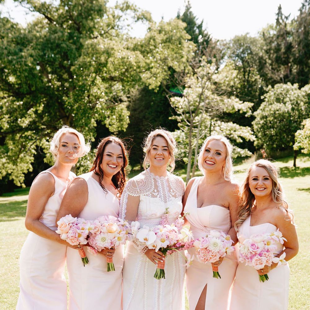 Bridesmaids in pink one shoulder bridesmaid dresses with bride in long sleeve lace wedding dress holding pink bouquets 