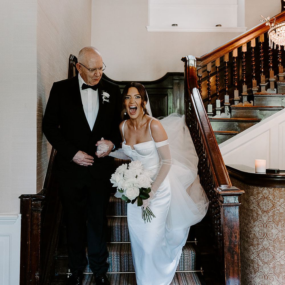 Father of the bride in black tuxedo walking with bride in satin wedding dress 