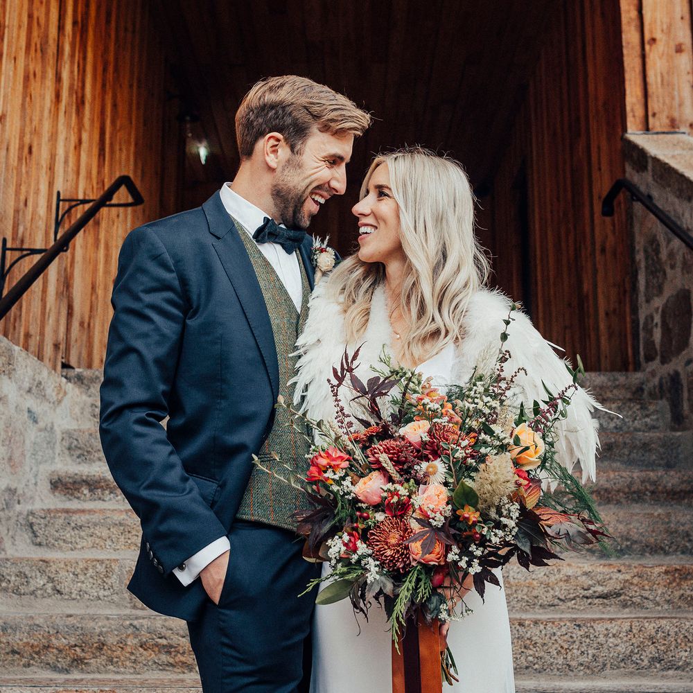Bride wearing feathered shawl and holding bouquet looks at groom on steps