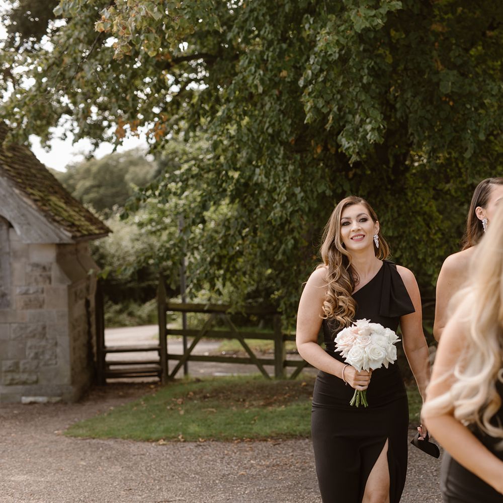 Bridesmaids in full length black dresses with long curled hair and white and blush pink rose bouquets walking outside church
