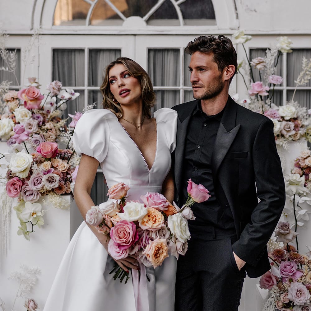 Groom in a black open shirt and tuxedo embracing his bride in a taffeta wedding dress as she holds a pink wedding bouquet 