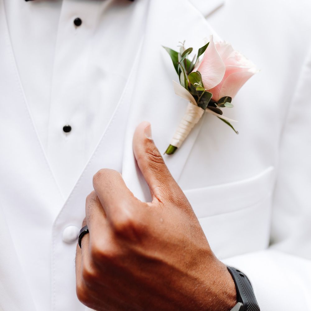 Groom in white shirt, white tuxedo jacket and black bow tie holds lapel of jacket next to pink rose buttonhole at backyard wedding