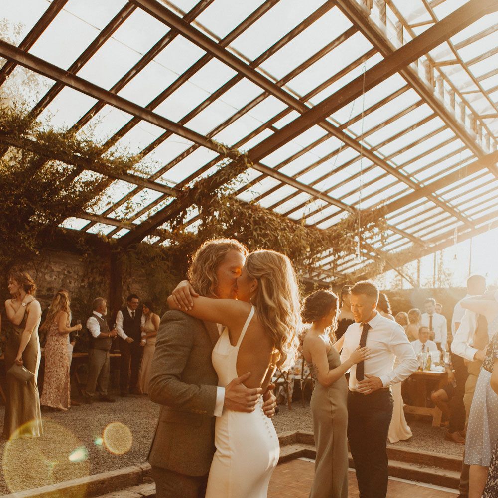 Bride in white open back Made With Love wedding dress with button details kisses groom in sage suit and brown shoes as they dance during golden hour at the glasshouse at Anran Devon