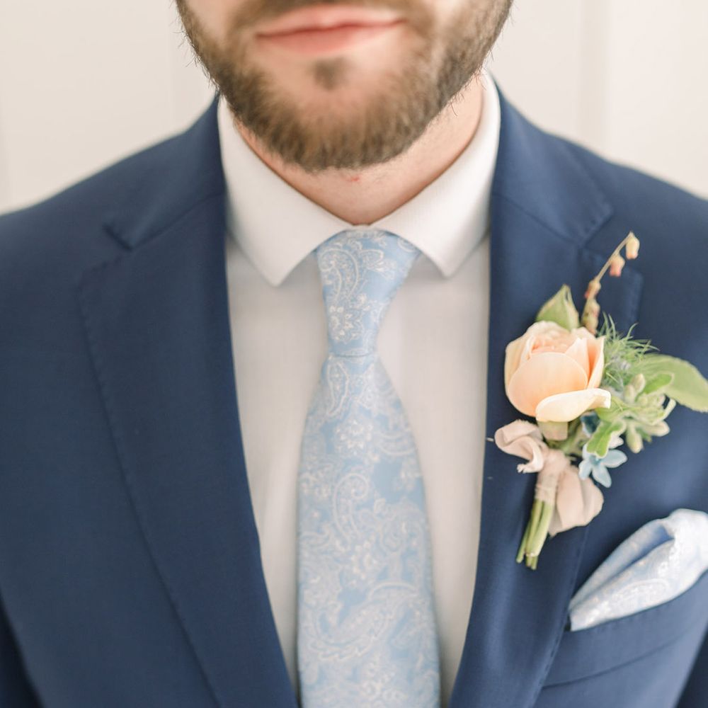 Groom in a navy jacket with white shirt, pale blue paisley tie and pocket square and peach rose buttonhole flower