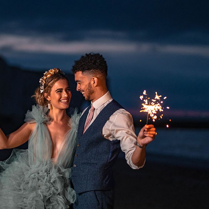 Bride in a mint green frilly wedding dress and groom a waistcoat holding sparklers on the beach 