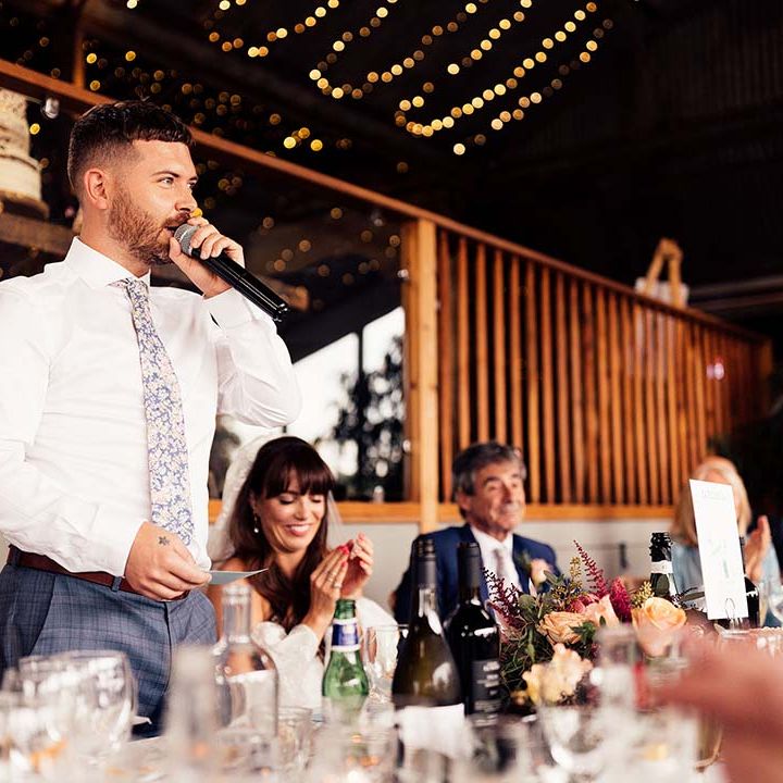 Groom in blue check trousers and a floral tie giving his wedding speech at Stone Barn Cotswolds wedding venue 