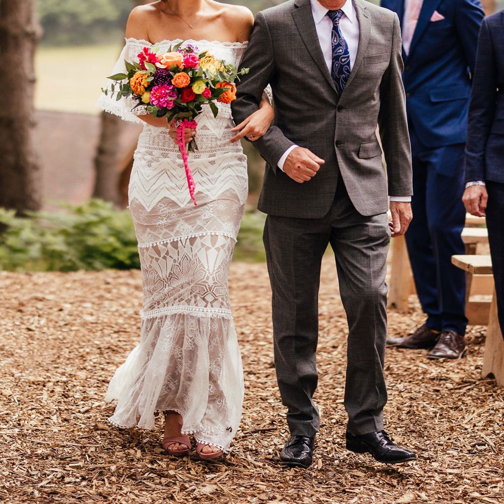 Bride in lace off the shoulder Grace Loves Lace wedding dress and flower crown carrying colourful wedding bouquet smiles whilst walking down outdoor aisle arm in arm with father of the bride in grey suit