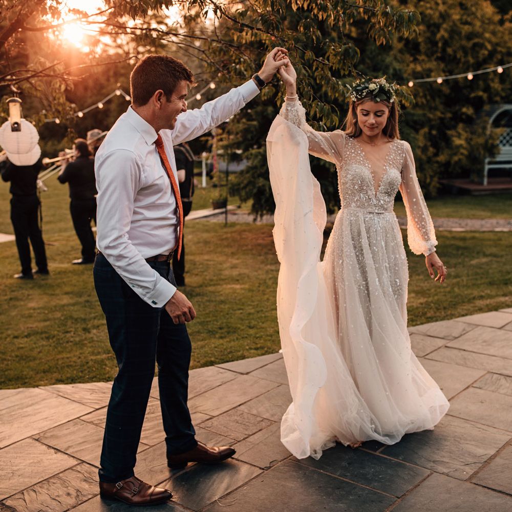 Bride in embroidered Julie Vino wedding dress and flower crown dances at golden hour with groom in white shirt at outdoor wedding reception 