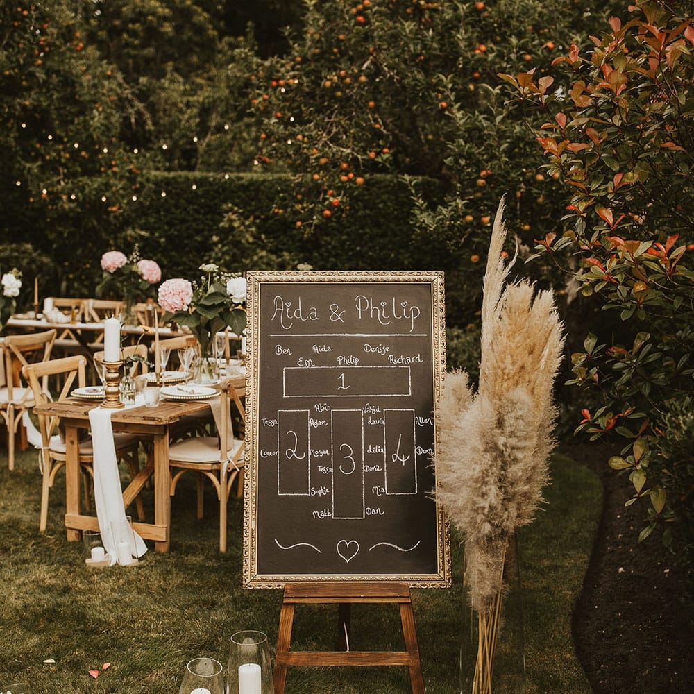 Chalkboard table plan within gold frame, on wooden easel next to pampas grass bouquet