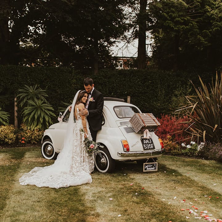 Bride & groom stand on lawn next to their restored Fiat 500 wedding car