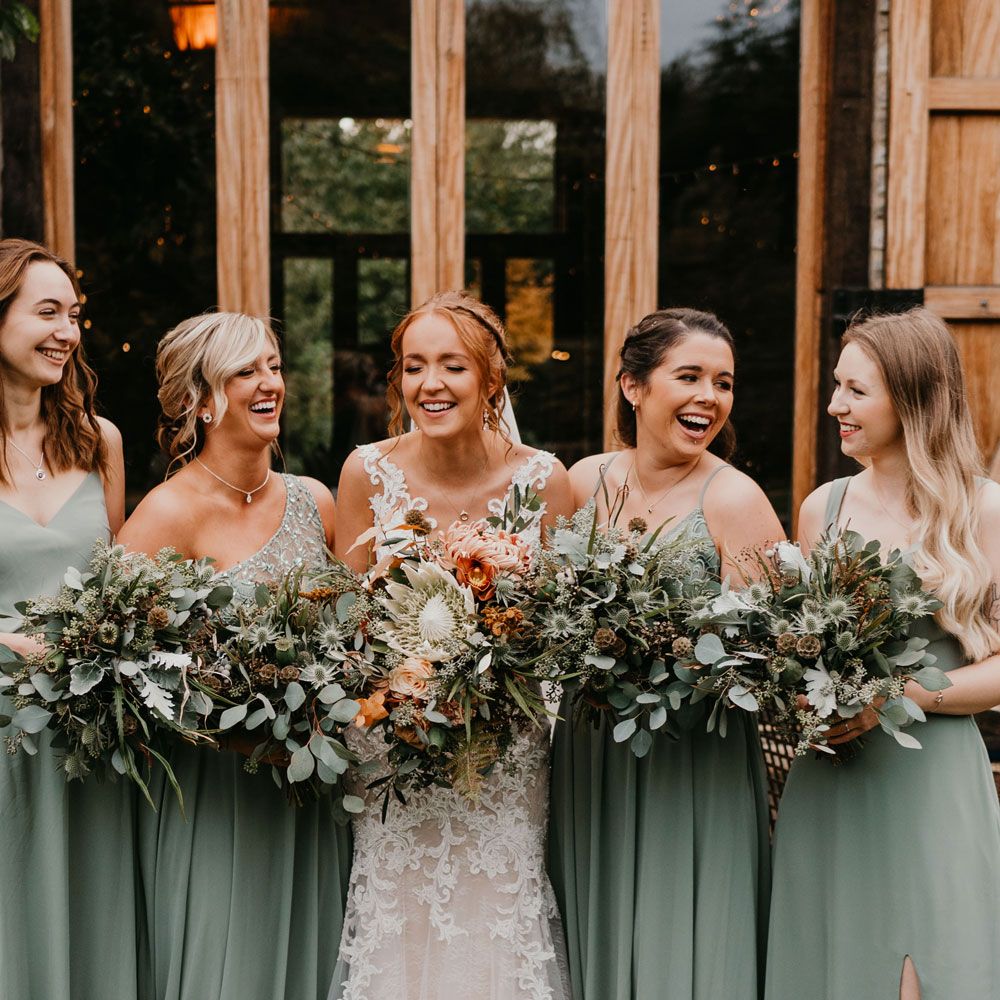 Bride with braided updo wearing lace wedding dress holding mixed bridal bouquet stands with bridesmaids in mismatched sage bridesmaid dresses holding green bouquets 