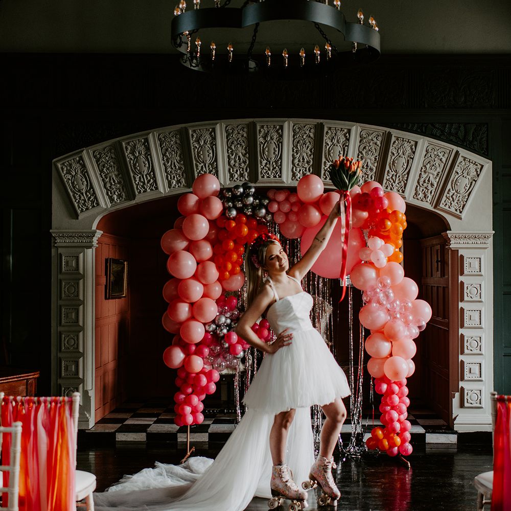 Bride in a short wedding dress wearing roller skates holding a tulip bouquet in the air at her fun, retro wedding with balloon installation