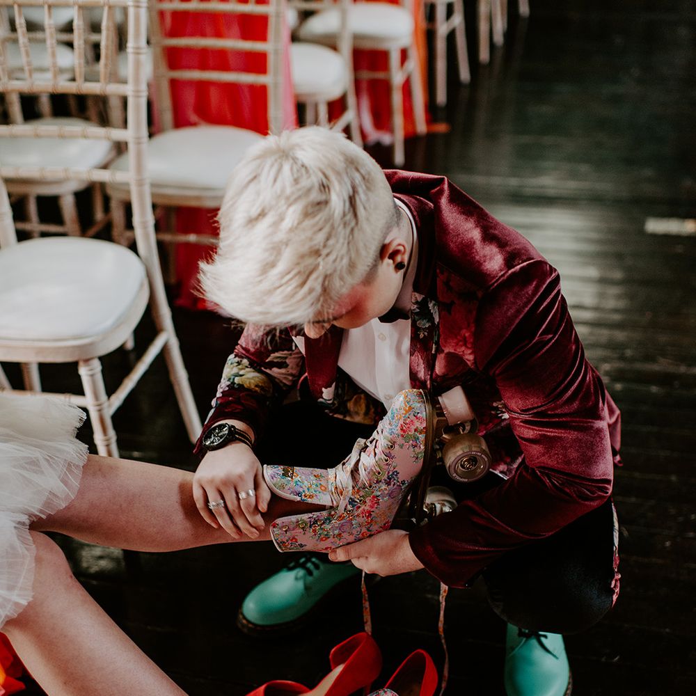 Bride with short platinum blonde hair and a burgundy velvet jacket putting on her brides floral retro roller skates 