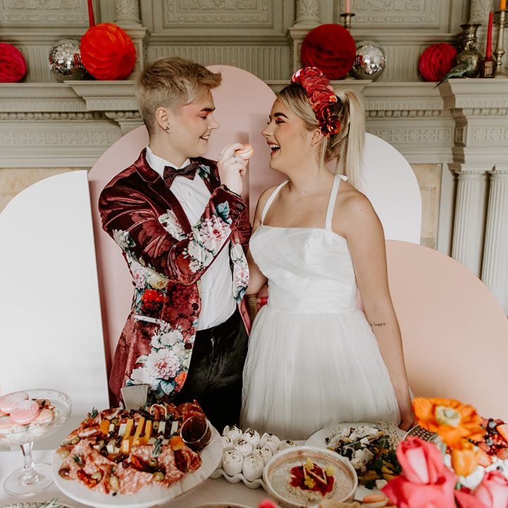 Two brides at the food table with pink geometric backdrop, velvet jacket and tulle skirt wedding dress 