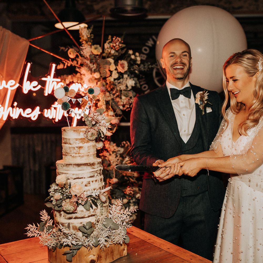 Groom in navy tweed suit with bow tie and buttonhole stands with bride in white wedding dress with pearl mesh overlay as she cuts three tier semi naked wedding cake