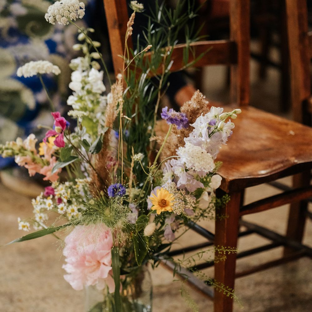 Colourful wildflower wedding flower displays in glass vases for aisle decoration next to dark wooden chairs