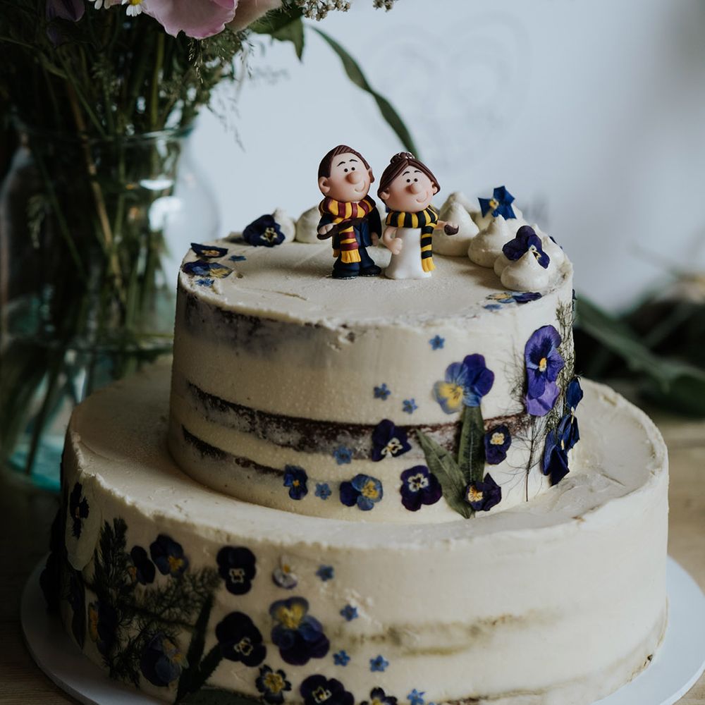 Semi naked two tier wedding cake with white frosting and edible flowers topped with custom bride and groom cake topper in Harry Potter style scarves 