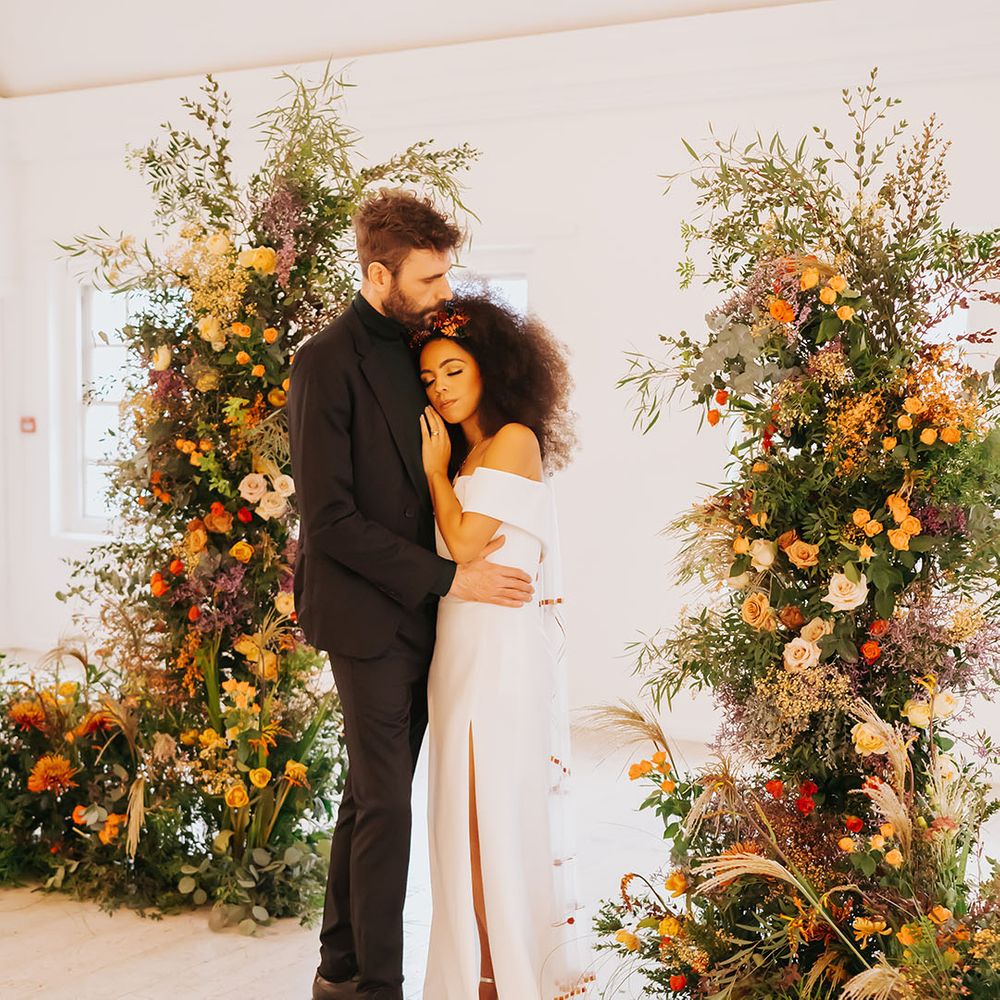 Black bride in an off the shoulder wedding dress with side slit and groom in all black embracing between column golden wedding ceremony flowers