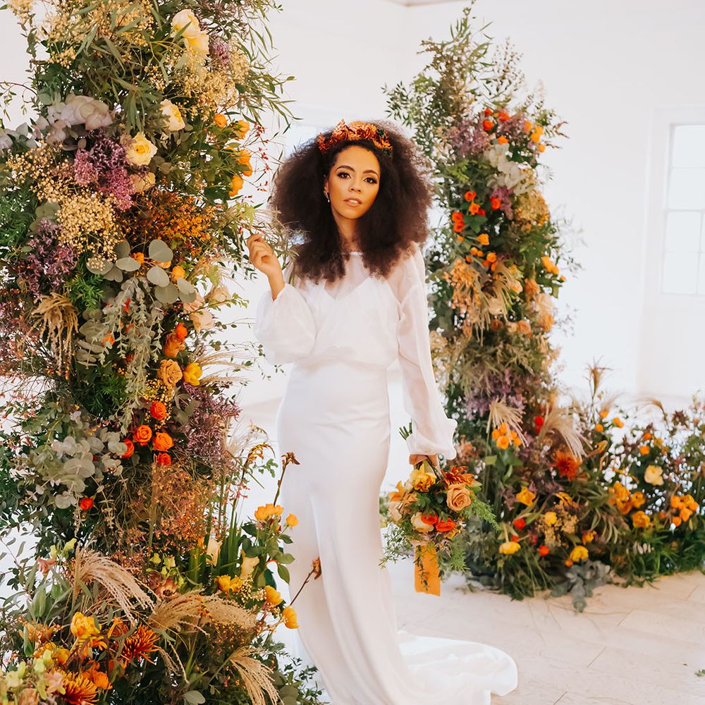 Black bride in a satin slip wedding dress with sheer top standing next to golden wedding, column flower arrangements 