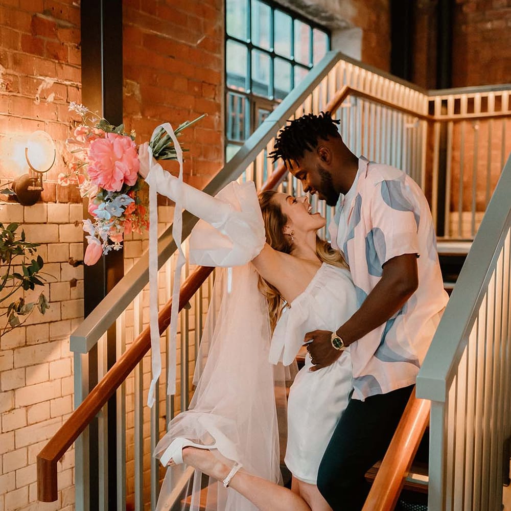 Groom in a pastel patterned shirt and Converse trainers embracing his bride in a short wedding dress with long veil holding her pink and blue bouquet in the air 