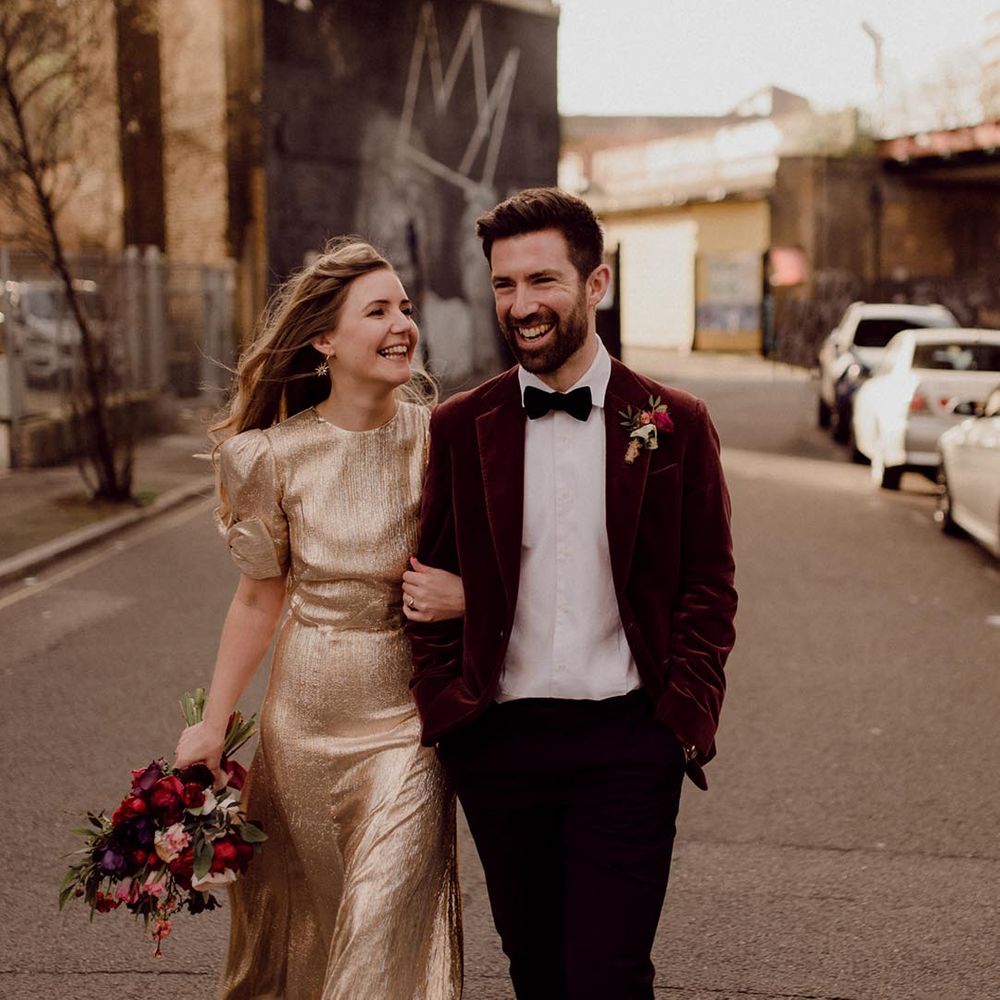 Bride in gold gown and star earrings links arms with groom in red velvet suit jacket at 100 Barrington city wedding venue