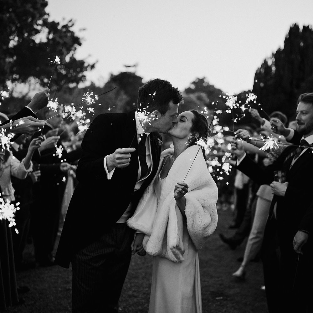 Bride and groom kiss as they wave sparkles and their guests wave them too with bride wearing white fur cover up 