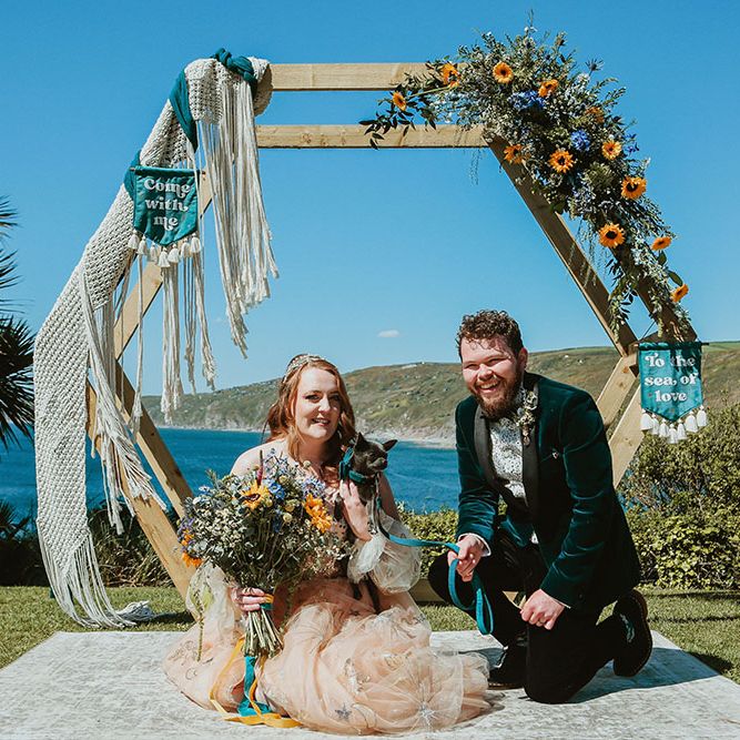 Bride & groom kneel with their dog in front of DIY wedding arch adorned with sunflowers and green foliage alongside blue fabric wedding banners