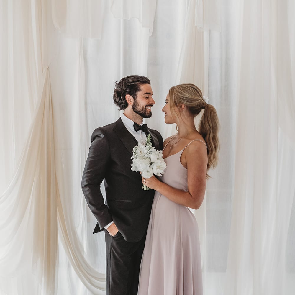 Groom in black tie with slicked back hair and a beard with bridesmaid in pastel dress with wavy ponytail hairstyle 
