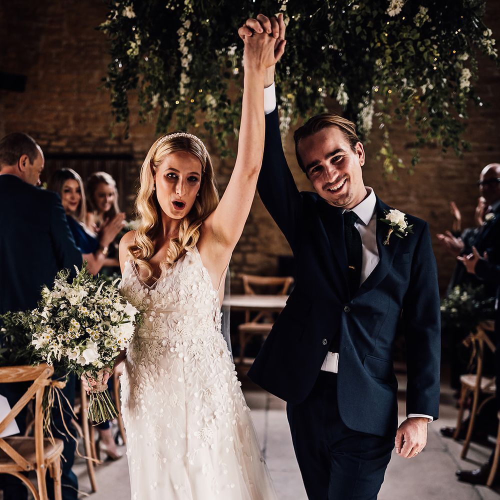 Groom in a navy blue suit with a white rose buttonhole walks back down the aisle with the bride 