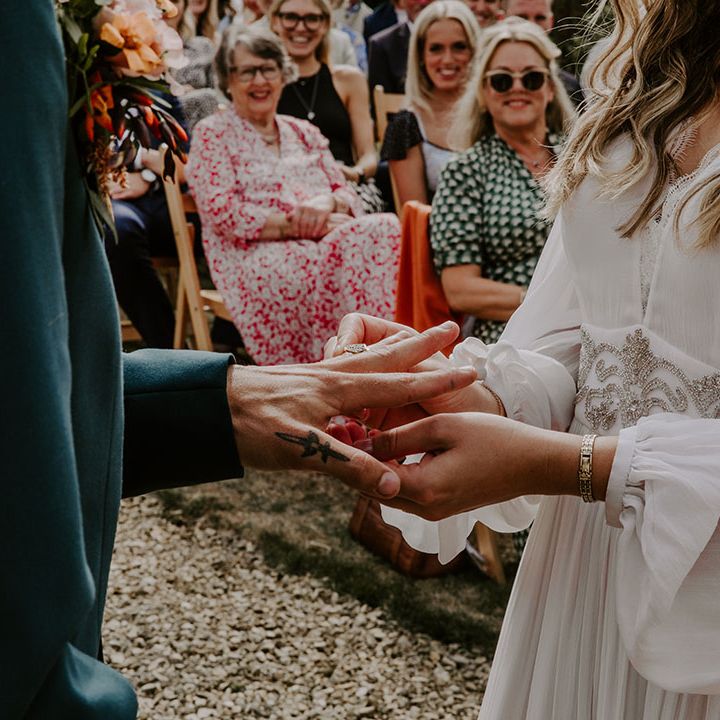 Bride places ring on her grooms finger as they stand outdoors during wedding ceremony 