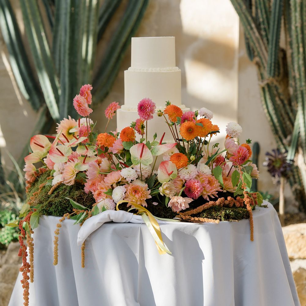 Brightly coloured floral arrangement surrounds four tier white frosted wedding cake 