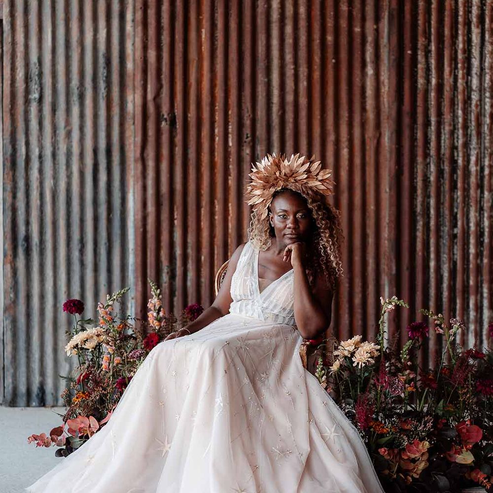 Bride in gold bridal crown, v neck peach wedding dress with embroidered stars sitting on red velvet chair and large autumnal dried floral arrangements