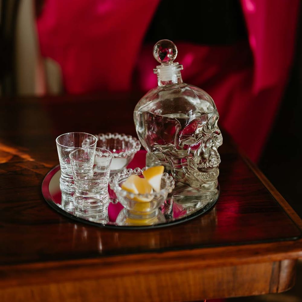 Skull shaped tequila decanter with two shot glasses, a small bowl of salt and a small bowl of limes on silver tray 