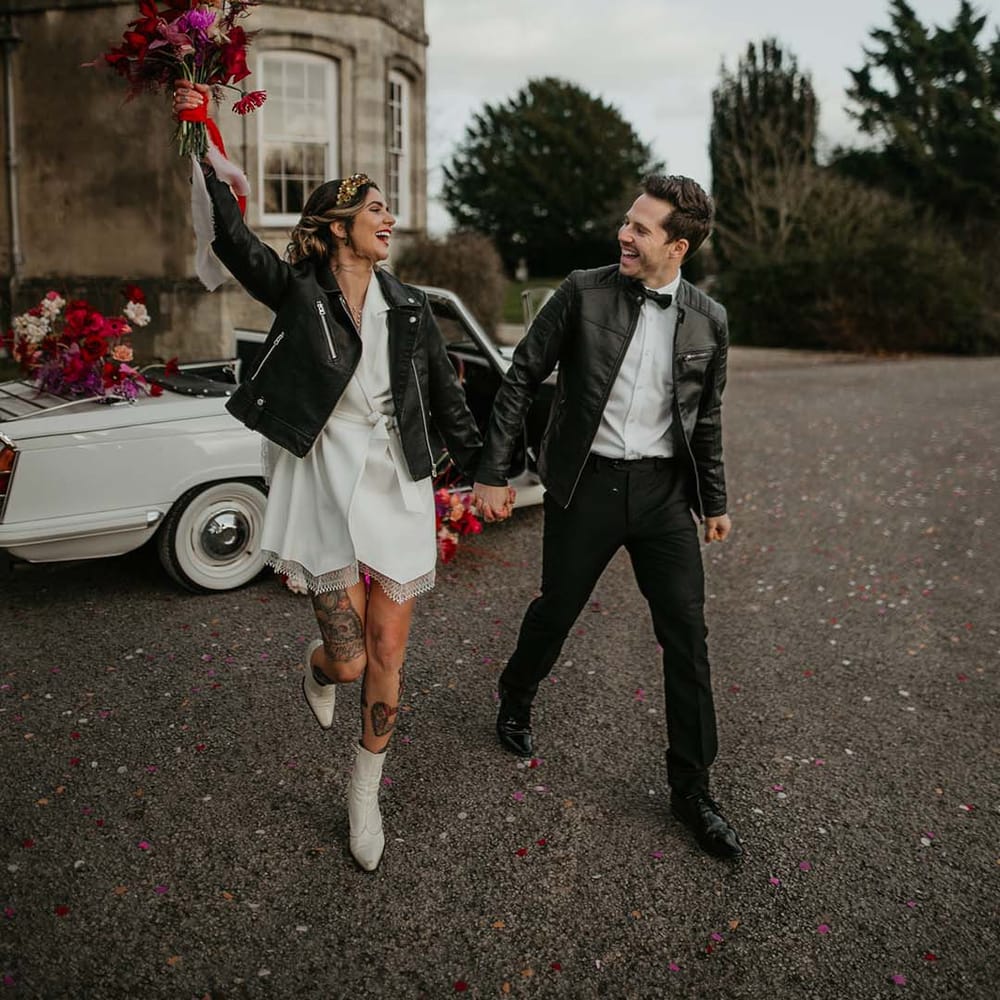 Groom in white shirt, black bowtie, black suit trousers and slim fitting black leather jacket and bride in blazer style wrap around short wedding dress with delicate fringe detailing and personalised, embroidered black leather jacket walking around the grounds of Elmore Court wedding venue