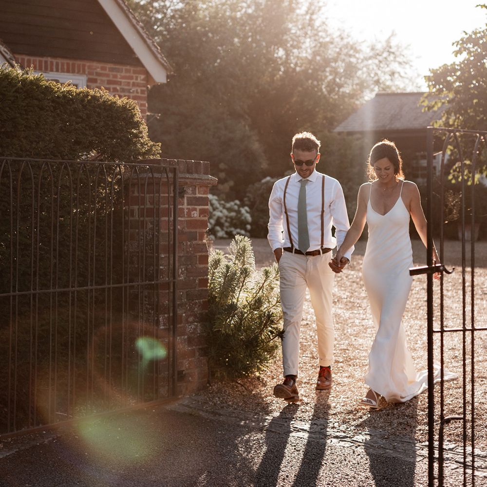 Groom in white shirt and sage green tie holds his brides hand as they walk outdoors during golden hour